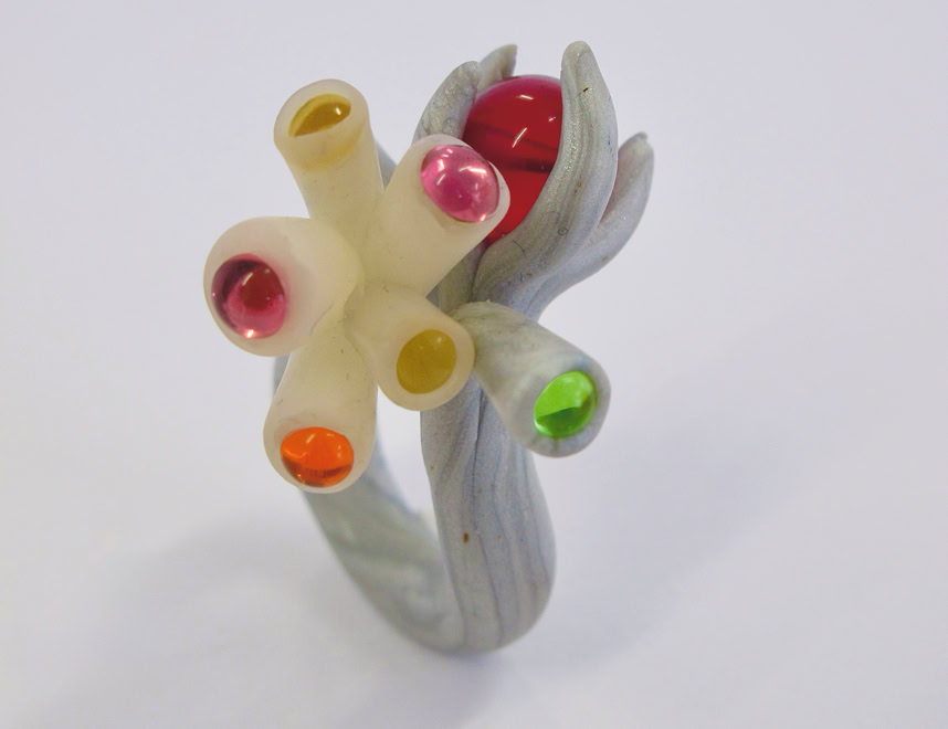 Bague sculpturale avec des tubes colorés et des sphères sur un fond blanc.