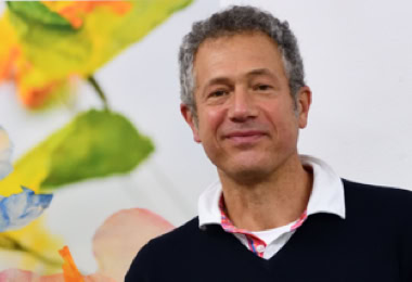 Photographie portrait de Denis Tcheskiss, par Georges Henz,  souriant portant un pull noir avec un col blanc, debout devant un fond coloré avec des motifs floraux flous.
