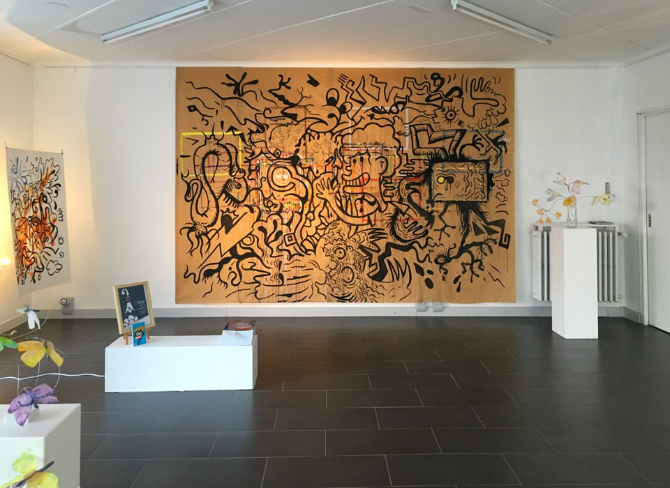 Salle d'exposition avec une grande peinture murale abstraite, des sculptures sur des socles blancs et un sol en carrelage sombre.