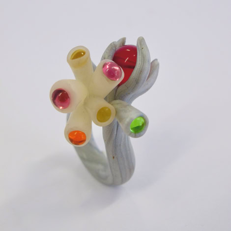 Bague sculpturale avec des tubes colorés et des sphères sur un fond blanc.
