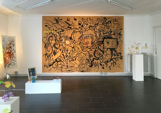 Salle d'exposition avec une grande peinture murale abstraite, des sculptures sur des socles blancs et un sol en carrelage sombre.