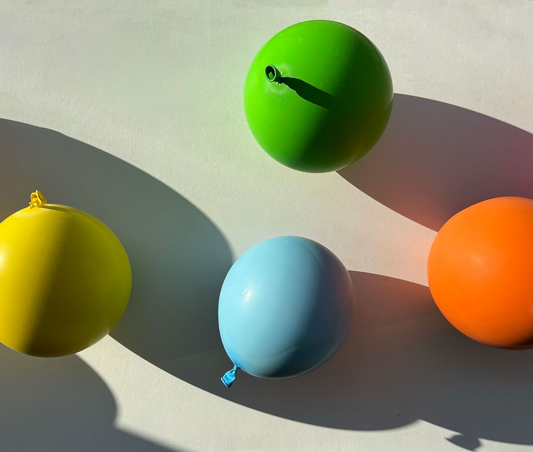 Quatre ballons colorés (jaune, vert, bleu, orange) sur une surface blanche avec des ombres.