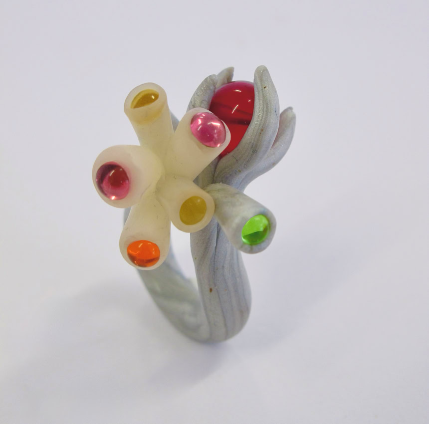 Bague sculpturale avec des tubes colorés et des sphères sur un fond blanc.