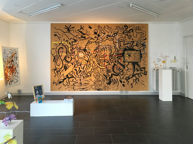 Salle d'exposition avec une grande peinture murale abstraite, des sculptures sur des socles blancs et un sol en carrelage sombre.