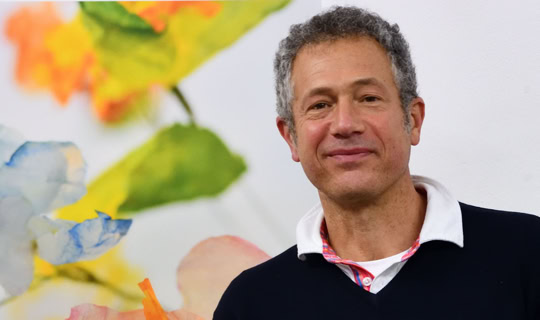 Photographie portrait de Denis Tcheskiss, par Georges Henz,  souriant portant un pull noir avec un col blanc, debout devant un fond coloré avec des motifs floraux flous.